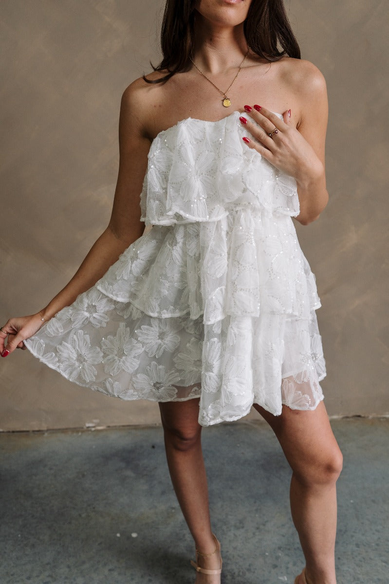 front view of model wearing the Kensley White Floral & Sequin Strapless Mini Dress that has white sheer fabric, a tiered body, a floral print outlined with sequins, and a sweetheart strapless neck.