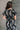 Back view of model wearing the Genevieve Black & Cream Printed Short Sleeve Top that has black fabric with a cream aztec pattern, a cropped waist, bubble hem, round neckline, a back key hole with a button, and short sleeves.