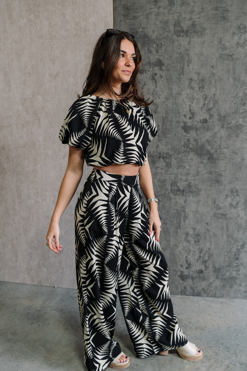 Full body frontal side view of model wearing the Genevieve Black & Cream Printed Wide Leg Pants that have black lightweight fabric with a cream aztec pattern, two side slit pockets, wide legs and an elastic waistband.