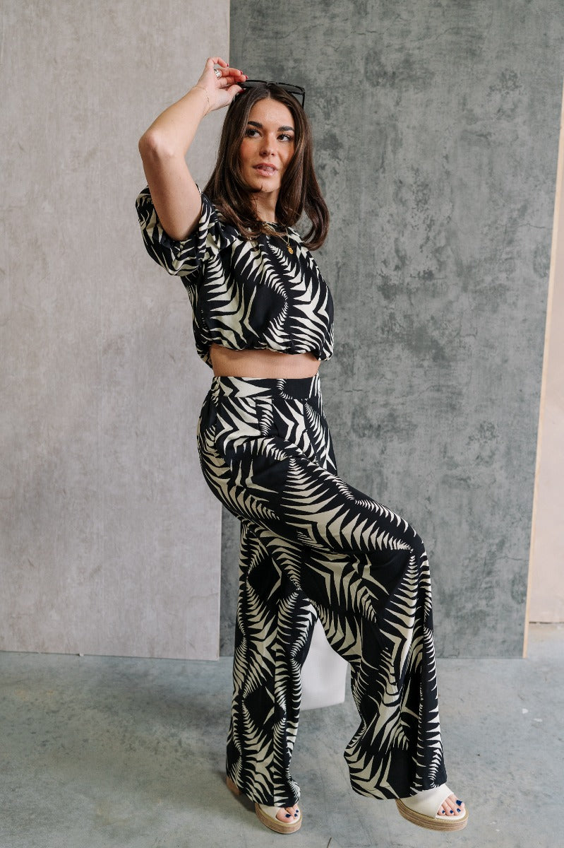 Full body side view of model wearing the Genevieve Black & Cream Printed Wide Leg Pants that have black lightweight fabric with a cream aztec pattern, two side slit pockets, wide legs and an elastic waistband.
