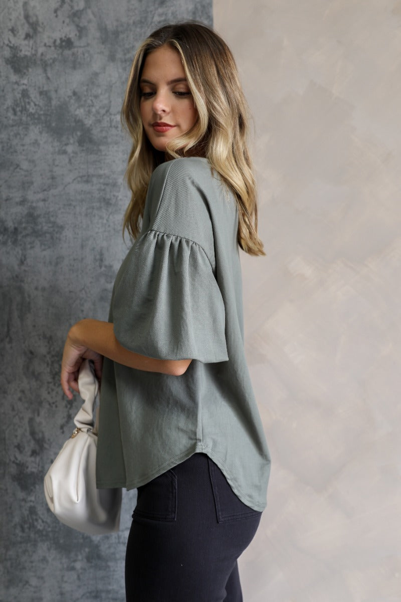 Side view ofo model wearing the Hazel Green Short Puff Sleeve Top which features light olive knit fabric, scooped hem, round neckline and short puff sleeves.
