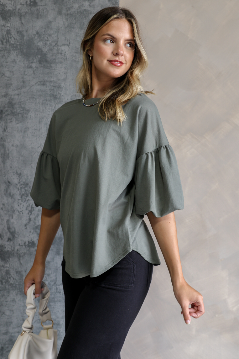 Side view ofo model wearing the Hazel Green Short Puff Sleeve Top which features light olive knit fabric, scooped hem, round neckline and short puff sleeves.