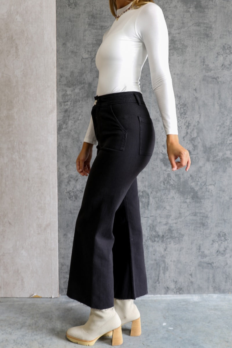 Side view of model wearing the Alexa Black High-Waisted Wide Leg Pants which features black denim fabric, two front pockets, two back pockets, front zipper with button closure, belt loops and wide pant legs.