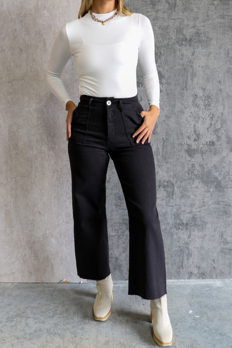 Front view of model wearing the Alexa Black High-Waisted Wide Leg Pants which features black denim fabric, two front pockets, two back pockets, front zipper with button closure, belt loops and wide pant legs.