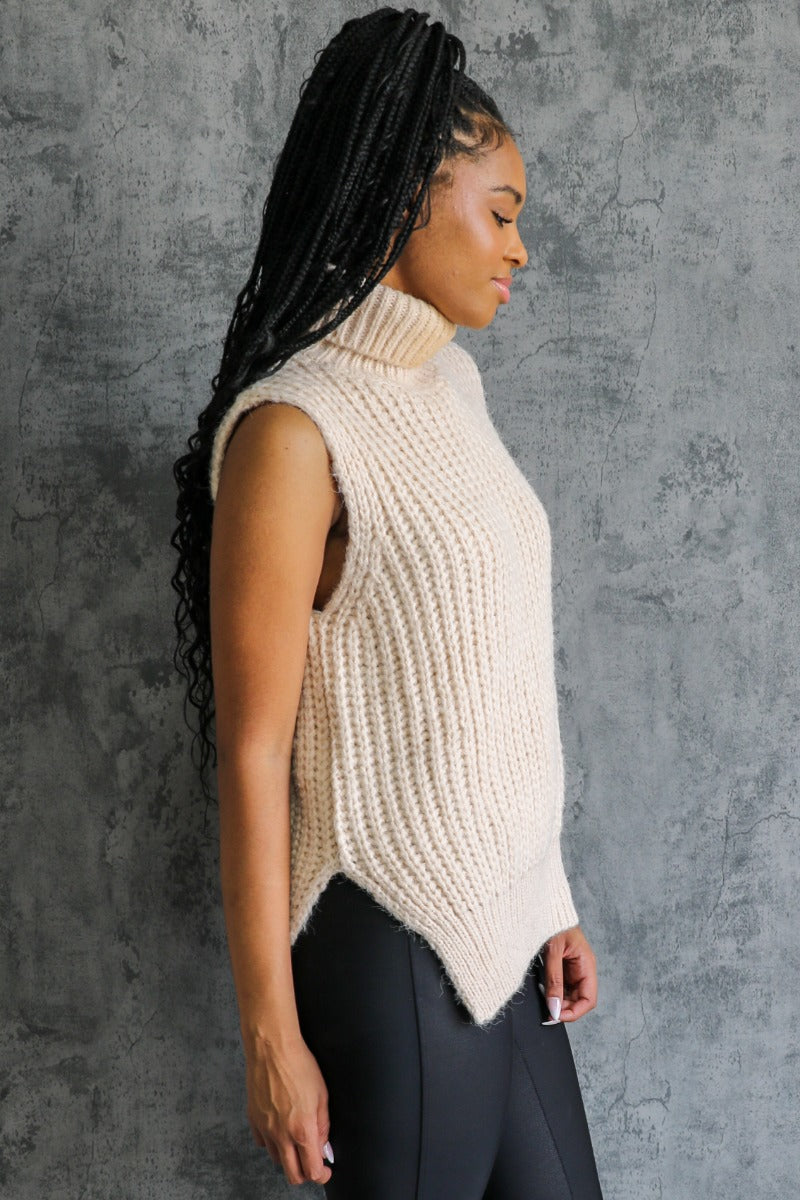 Side view of model wearing the Olivia Beige Cable Knit Turtleneck Sleeveless Sweater which features beige cable knit fabric, high low hem, turtleneck neckline and sleeveless.