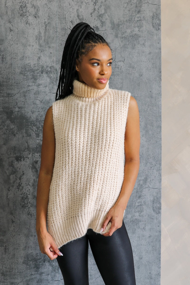Front view of model wearing the Olivia Beige Cable Knit Turtleneck Sleeveless Sweater which features beige cable knit fabric, high low hem, turtleneck neckline and sleeveless.