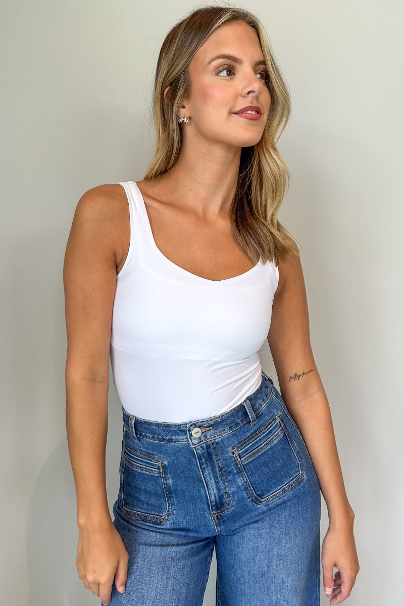 Front view of model wearing the Avery White Sleeveless Bodysuit which features white knit fabric, a scooped neckline, thick straps, and a thong bottom with snap closures.