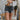 Front view of model wearing the The Rooted Denim: Take It Easy Shorts that have charcoal washed denim, two front pockets, a distressed hem, belt loops , and back pockets.