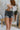 Front view of model wearing the The Rooted Denim: Take It Easy Shorts that have charcoal washed denim, two front pockets, a distressed hem, belt loops , and back pockets.