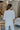 Back view of model wearing the Simply Yours Top that has ivory fabric with tan stitched stripes, a high-low hem, a notched neck with a tie and 3/4 sleeves