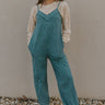 Full body front view of model wearing the Ready To Road Trip Jumpsuit that has washed blue fabric, a v-neck, distressing, two side pockets, thin straps, and wide pants legs with elastic hems.