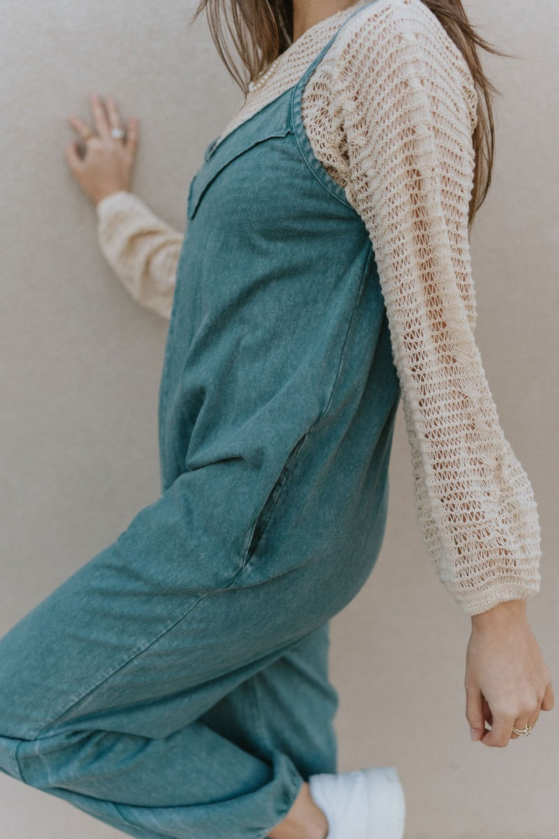 Upper side view of model wearing the Ready To Road Trip Jumpsuit that has washed blue fabric, a v-neck, distressing, two side pockets, thin straps, and wide pants legs with elastic hems.