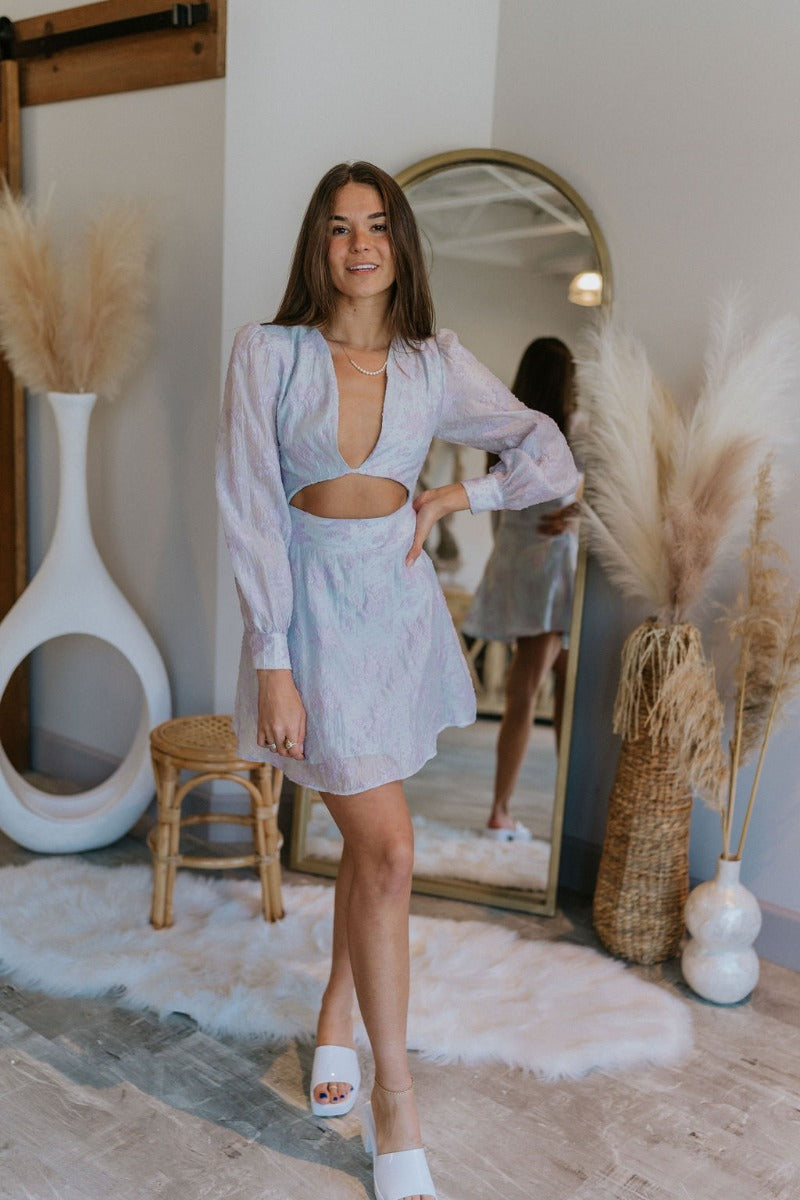 Full body view of model wearing the Enchanted Times Dress which features light blue and light purple fabric with purple embroidered flowers, a lining, a mini-length hem, a cutout in the mid section, a plunge neckline, a back zipper, and sheer balloon slee