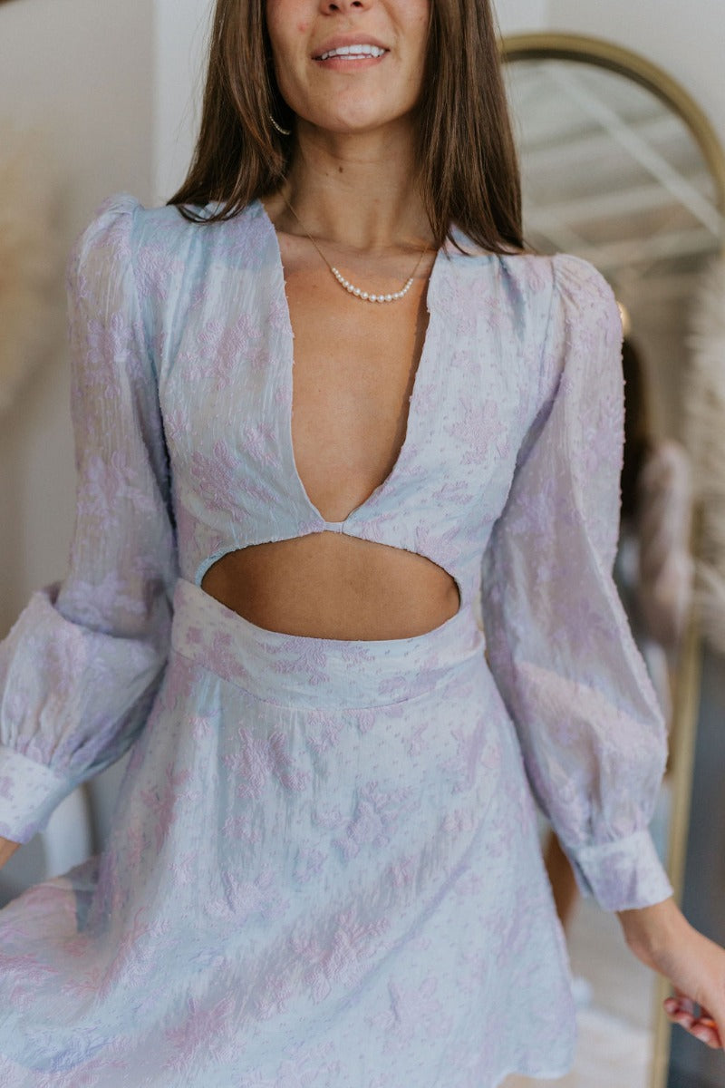 Close up view of model wearing the Enchanted Times Dress which features light blue and light purple fabric with purple embroidered flowers, a lining, a mini-length hem, a cutout in the mid section, a plunge neckline, a back zipper, and sheer balloon sleev