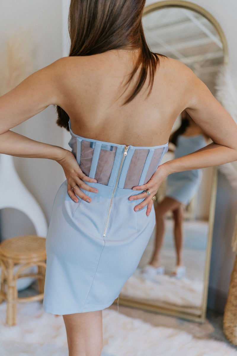 Close up/back view of model wearing the Wishful Thinking Dress in Blue which features light blue fabric, mini length, overlap neckline with extra fabric, back sheer fabric with bands and gold zipper.