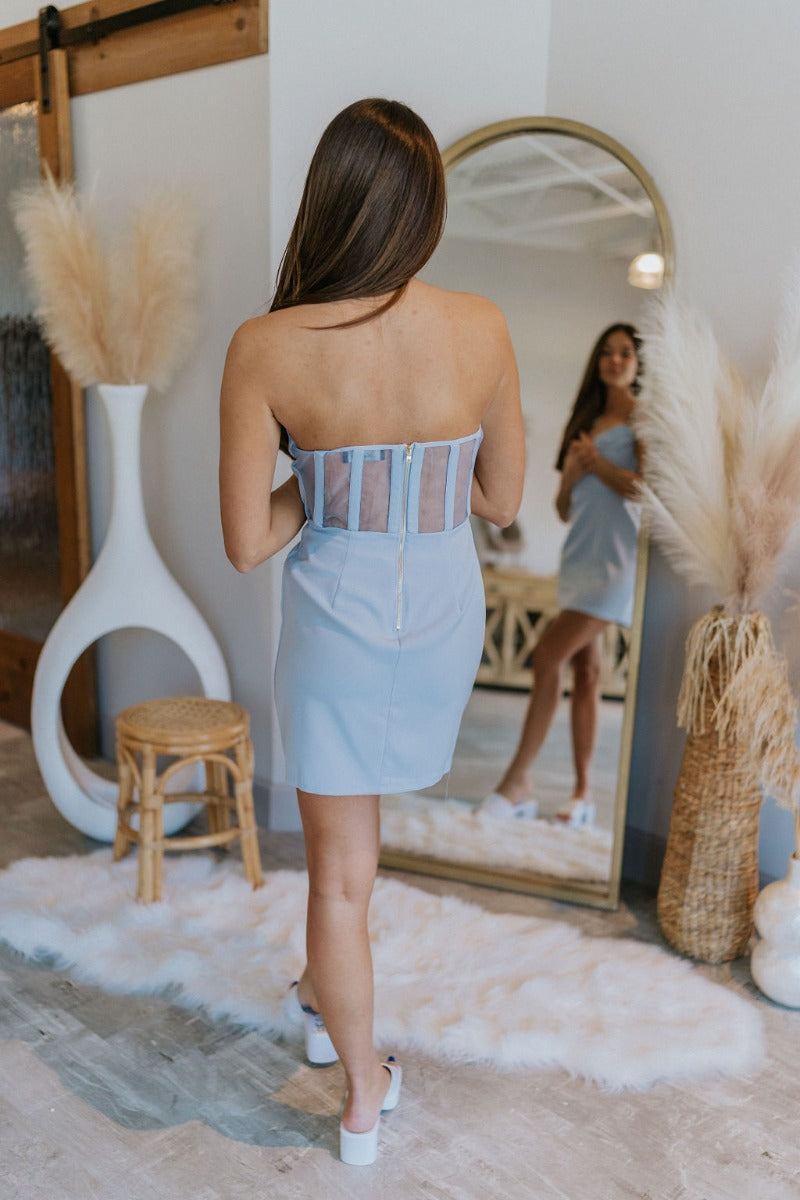 Full body/back view of model wearing the Wishful Thinking Dress in Blue which features light blue fabric, mini length, overlap neckline with extra fabric, back sheer fabric with bands and gold zipper.