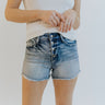 Front view of model wearing the Rooted Denim: Walk In The Park Shorts which features medium washed denim, two front pockets, a distressed hem, a 4-button fly with a zipper closure, belts loops on the waistband, and two back pockets.
