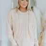 Close up view of model wearing the Now or Never Top which features light peach fabric, one front pocket, a high-low hem with slits on the side, distressed details, a v-neckline, a hood with drawstrings, dropped shoulders, and long sleeves with cuffs.