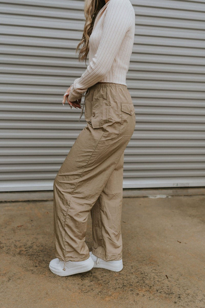 Side view of model wearing the Nothing On You Pants which features taupe nylon fabric, front buttoned pockets, back buttoned pockets, an elastic waistband with drawstring ties, and wide legs with drawstring hems.