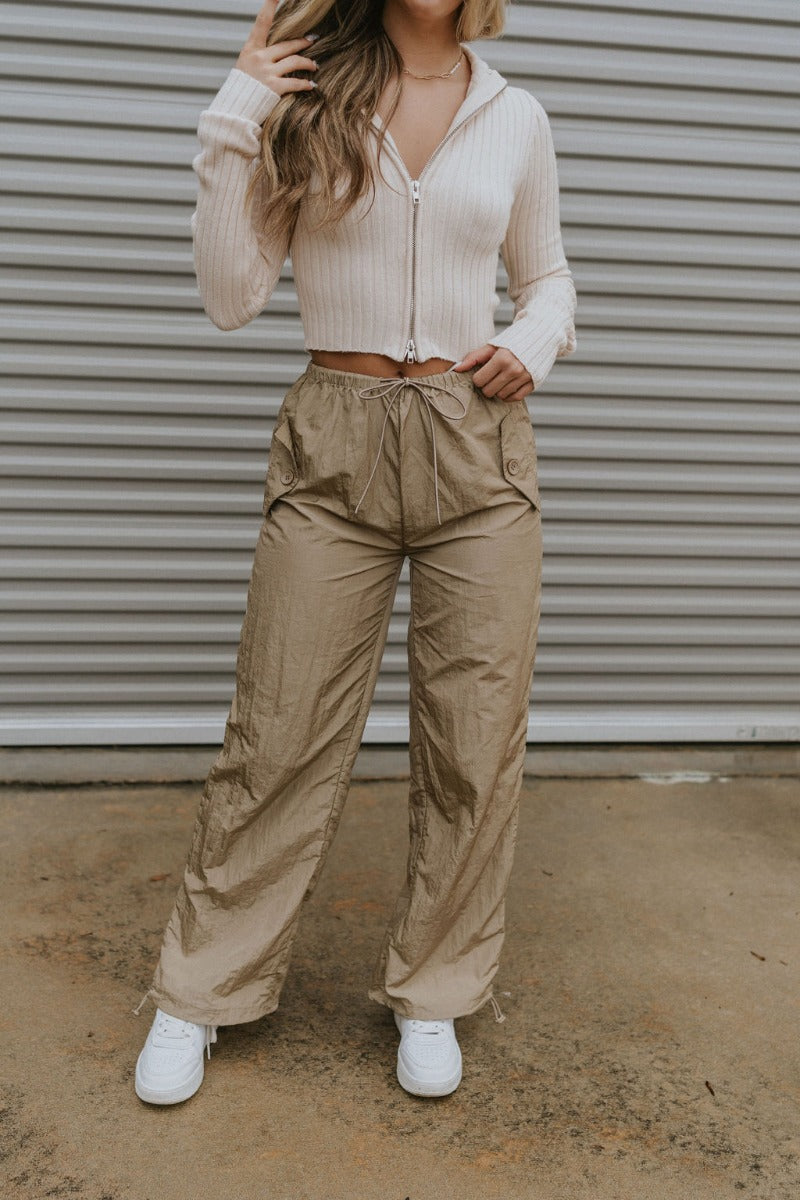 Front view of model wearing the Nothing On You Pants which features taupe nylon fabric, front buttoned pockets, back buttoned pockets, an elastic waistband with drawstring ties, and wide legs with drawstring hems.