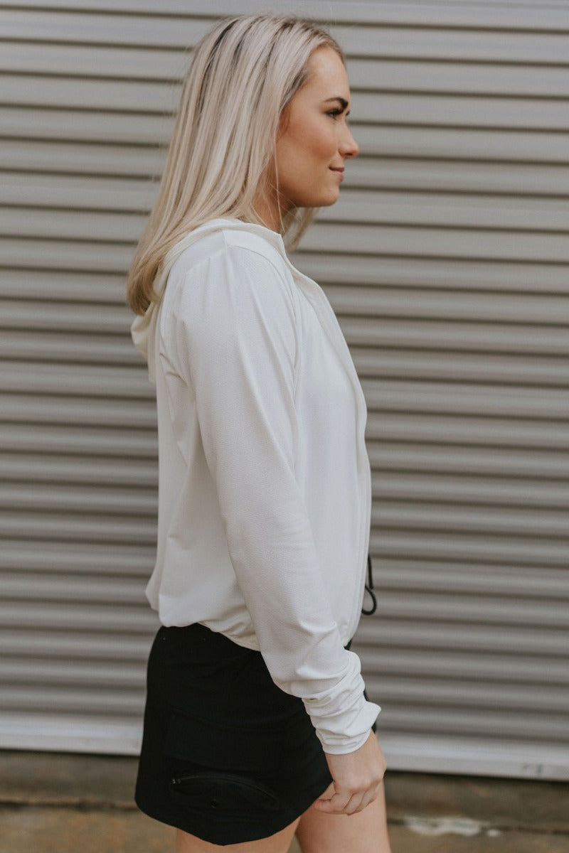 Side view of model wearing the All I Want Lightweight Jacket features white fabric, a monochromatic zip up, two front pockets, a high neckline with drawstrings and a hood, an elastic hem, a lining in the front, and long sleeves with cuffs.