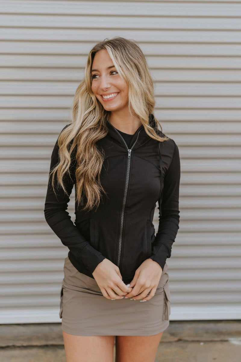 Front view of model wearing the Working It Jacket in Black which features black athleisure fabric, a monochromatic zip up, two front pockets, a high neckline with drawstrings and a hoodie attached, a lining, and long sleeves.