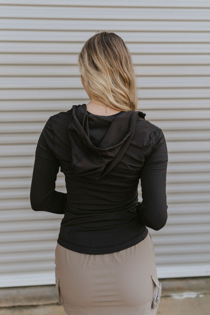 Back view of model wearing the Working It Jacket in Black which features black athleisure fabric, a monochromatic zip up, two front pockets, a high neckline with drawstrings and a hoodie attached, a lining, and long sleeves.