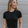 Close up view of model wearing the Raise The Standard Bodysuit in Black which features black color fabric, round neckline, short sleeves and thong bottom with button closures.