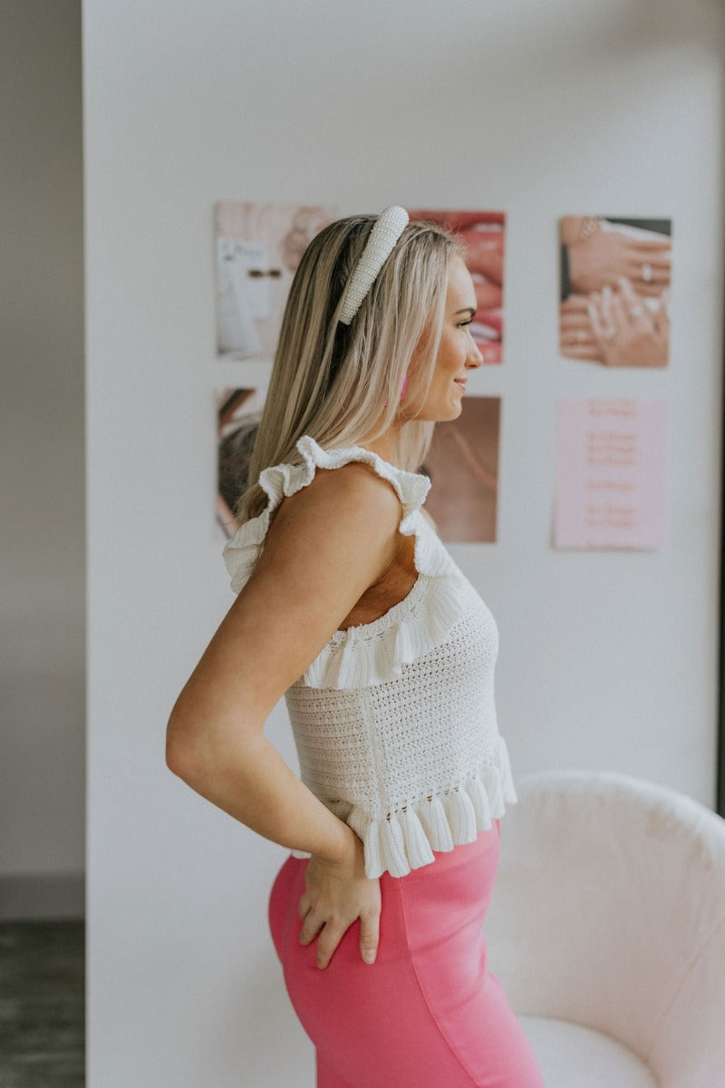 Side view of model wearing the All About You Top that has cream crochet fabric, a round neckline, a ruffle hem, a buttoned key hole in the back, and a sleeveless body with ruffles