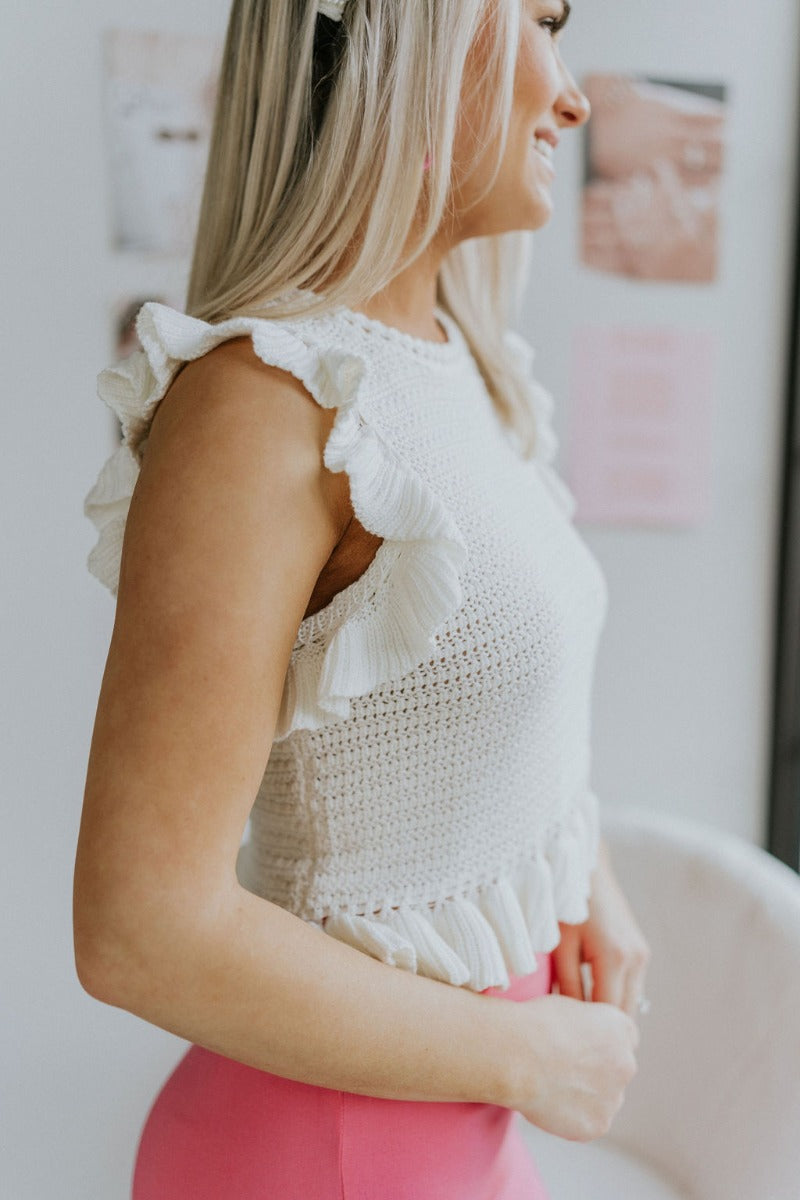 Side  view of model wearing the All About You Top that has cream crochet fabric, a round neckline, a ruffle hem, a buttoned key hole in the back, and a sleeveless body with ruffles