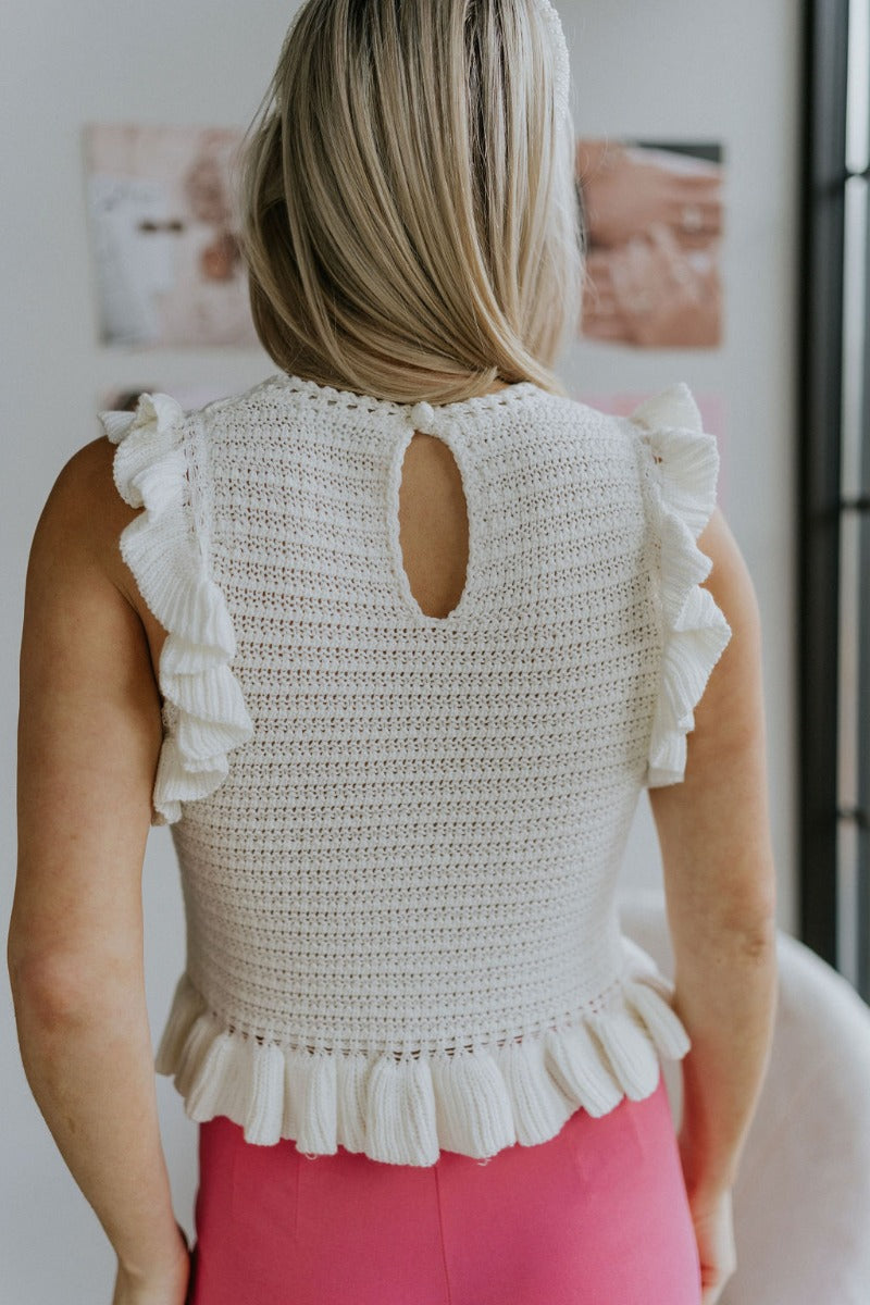 Back view of model wearing the All About You Top that has cream crochet fabric, a round neckline, a ruffle hem, a buttoned key hole in the back, and a sleeveless body with ruffles