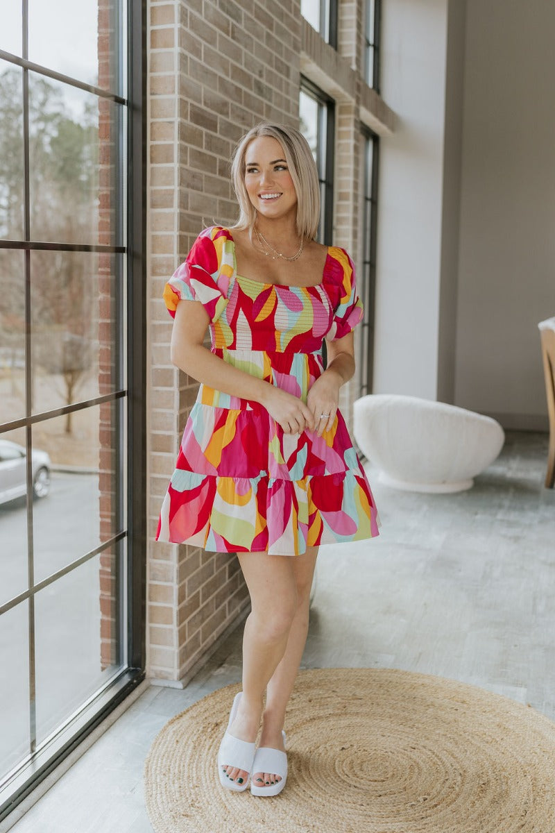 Full body view of model wearing the Colorful Dreams Dress which features red, pink, orange, lime green, aqua blue and cream fabric, geometric pattern, mini length, tiered style, smocked chest, square neckline and short puff sleeves.