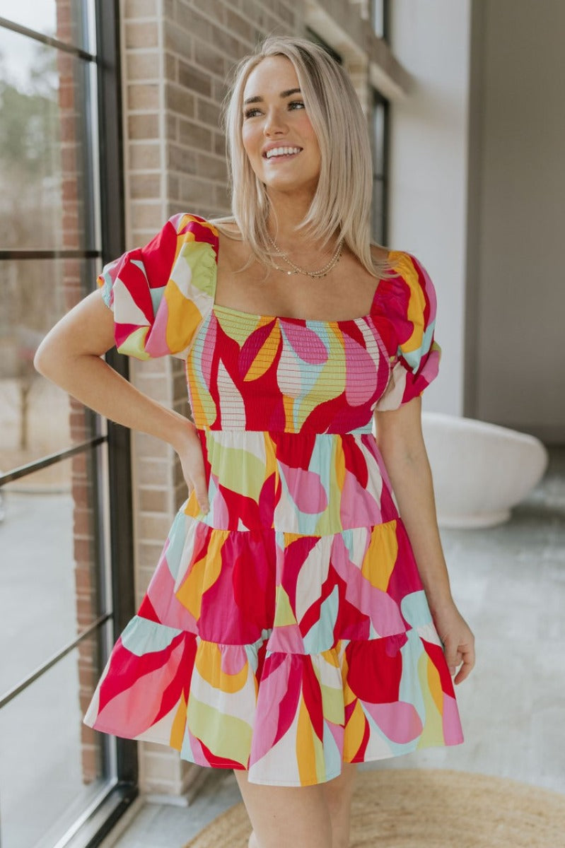 Front view of model wearing the Colorful Dreams Dress which features red, pink, orange, lime green, aqua blue and cream fabric, geometric pattern, mini length, tiered style, smocked chest, square neckline and short puff sleeves.