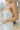 Close up/side view of model wearing the Wishful Thinking Dress which features white fabric, mini length, overlap neckline with extra fabric, back sheer fabric with bands and gold zipper.