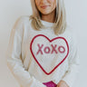 Close up view of model wearing the Love Always Sweater which features cream loose-knit fabric, a round neckline, and long sleeves with cuffs. The sweater says "XOXO" in light pink frill with a hot pink heart.