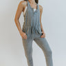 Front view of model wearing the Tell Me More Jumpsuit that has soft heather grey fabric, a quarter button-up neckline, side pockets, a drawstring around the waistline, a racer back, and jogger pants with thick hems