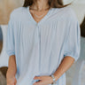 Close up view of model wearing the Take Me Away Top which features light blue fabric, a high-low hem, two pearlescent buttons with a v-neckline, and short puff sleeves with elastic bands.