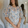 Front view of model wearing the Tea Party Blouse which features light blue net fabric, white tank lining, blue floral textured pattern with sequins, round neckline, back key hole with button closure and short puff sleeves.