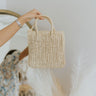 Close up view of model holding the Soak In The Sun Purse which features straw material with cream coloring, straw wrapped straps and a long braided strap and zipper closure.