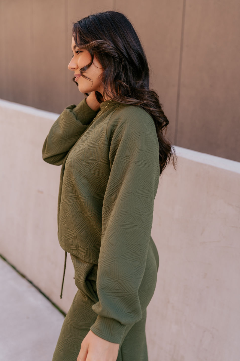 Side view of model wearing the Aria Olive Green Pattern Lounge Top which features olive green knit fabric, a monochrome geometric quilted pattern, a round neckline, and long balloon sleeves with cuffs.