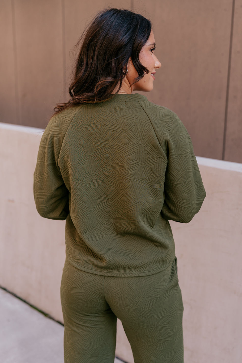 Back view of model wearing the Aria Olive Green Pattern Lounge Top which features olive green knit fabric, a monochrome geometric quilted pattern, a round neckline, and long balloon sleeves with cuffs.