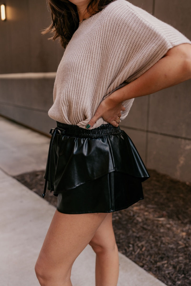 Side view of model wearing the Kali Black Faux Leather Ruffle Skort which features black faux leather, ruffle tier design, black shorts lining and elastic waistband with tie closure.