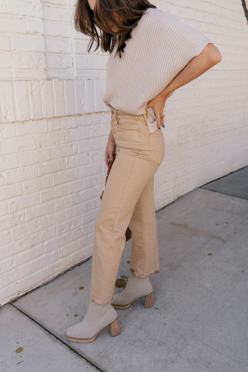Side view of model wearing the Rooted Denim: Morgan Light Mustard Straight Leg Jeans that have pale mustard/brown denim fabric,  pockets, and a frayed hem.