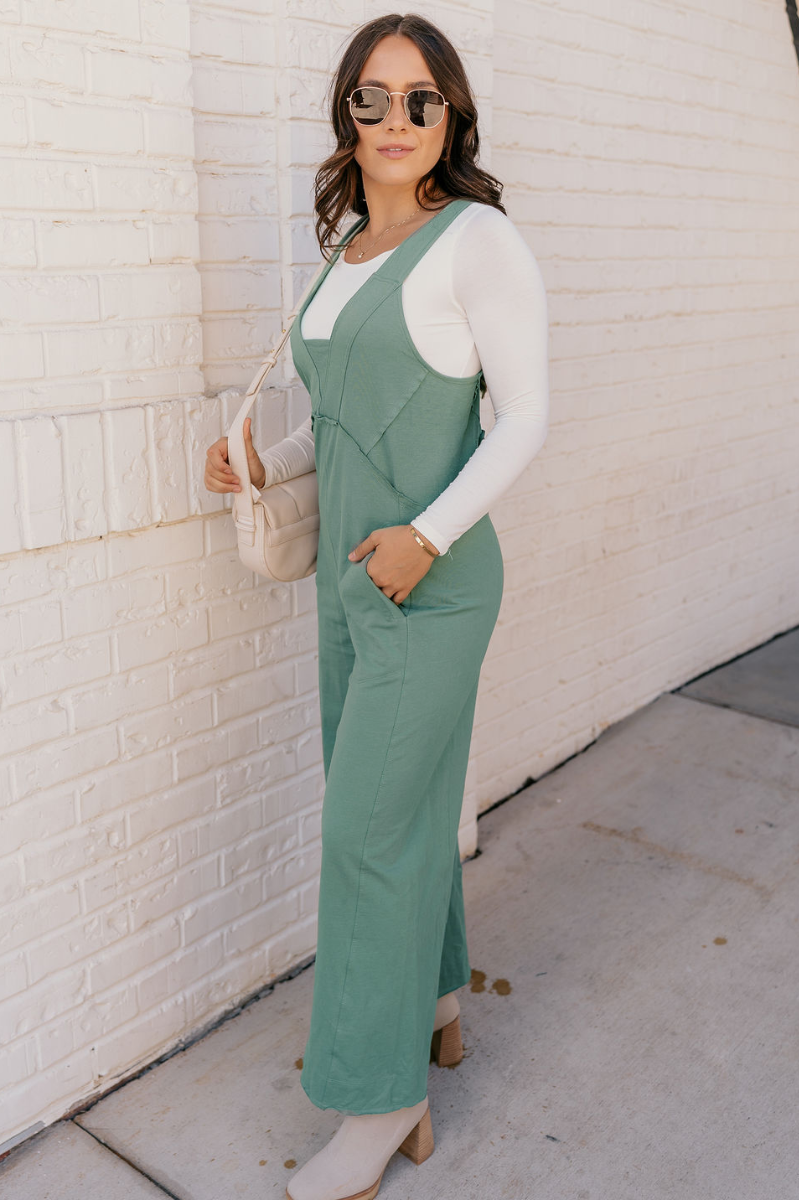 Full body view of model wearing the Lyla Sage Sleeveless Jumpsuit which features sage cotton fabric, raw hem details, two front pockets, a v-neckline, thick straps, a sleeveless design, and wide pant legs.