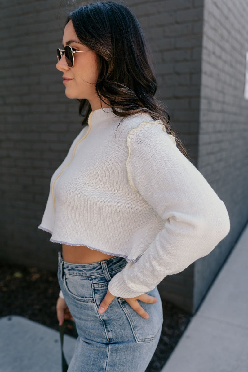 Side view of model wearing the Raegan Ivory Multi-Color Seam Sweater features white knit fabric with colorful exposed seam details, a high round neck, a cropped waist, and long sleeves.