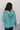 Back view of model wearing the Lottie Teal Green Cable Knit Hoodie Sweater that has teal knit fabric, a high-low hem, a hooded with black and cream ties, dropped shoulders, and long sleeves.