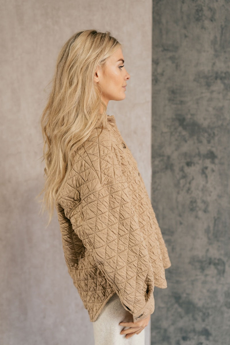 side view of model wearing the Kira Taupe Quilted Shacket that has taupe textured fabric with a quilted pattern, a left front chest pocket, a scooped hem, tortoise buttons, a collar, and long sleeves.