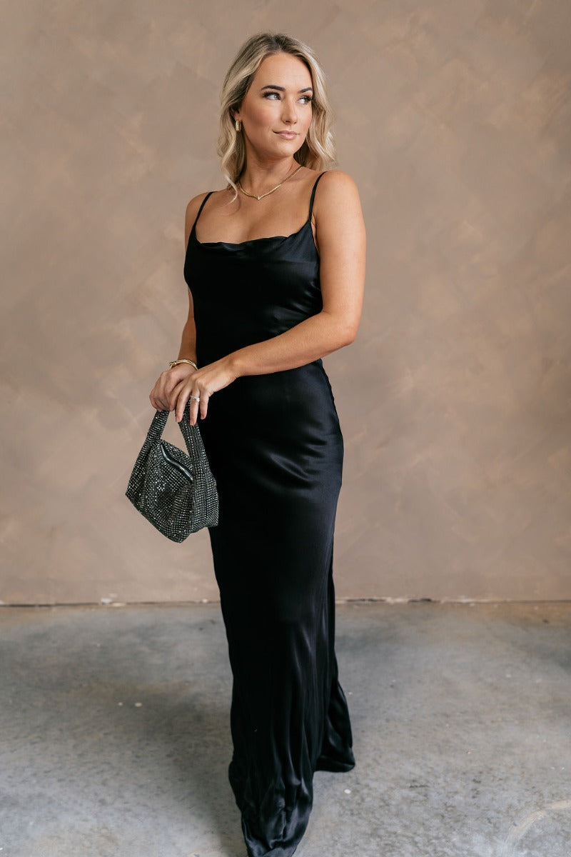 Full body front view of model wearing theElisa Black Satin Cowl Neck Maxi Dress that havs black satin fabric, maxi length,, a cowl neck, adjustable straps, and a back zipper with a hook closure.