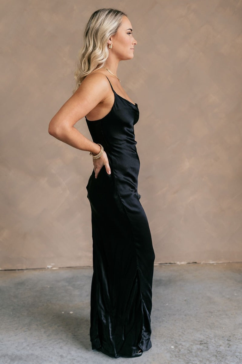 Full body side view of model wearing theElisa Black Satin Cowl Neck Maxi Dress that havs black satin fabric, maxi length,, a cowl neck, adjustable straps, and a back zipper with a hook closure.
