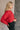 Side view of model wearing the Amara Red Cable Knit Long Sleeve Sweater which features red knit fabric, monochrome cable knit design, ribbed hem, cropped waist, round neckline, dropped shoulders and long sleeves with cuffs.